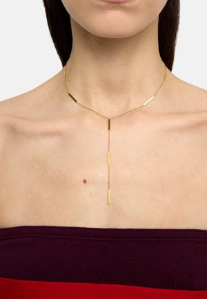 Złoty naszyjnik lariat z pięcioma pionowymi prostokątnymi zawieszkami noszony na nagiej szyi i ramionach, w parze z gorsetową bordowo-czerwoną bluzką.