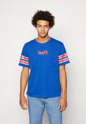 Camiseta de algodón azul con el logotipo rojo de "levi's" en el pecho, con mangas cortas a rayas blancas y rojas. Llevada con jeans de color azul claro.