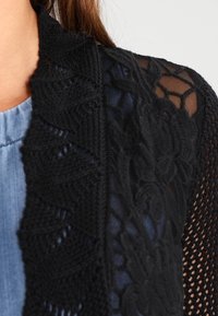 Svart spetskofta med en texturerad blommönster och transparenta ärmar, bärs över en ljusblå topp. Mjuk stickad material, öppen design.