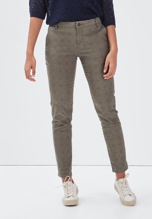 Žena v olivově zelených úzkých kalhotách s drobnými potisky, tmavě modrém svetru a bílých teniskách stojící před jednobarevným pozadím.