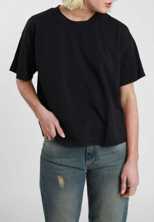 Persona bionda indossa una maglietta nera a maniche corte e jeans blu strappati, con una mano in tasca e la testa girata verso destra.