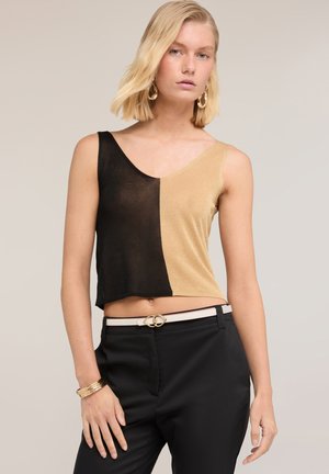 Kvinde iført ærmeløs, asymmetrisk sort og gylden top med sorte bukser, hvidt bælte, gyldne hoop øreringe og gyldne armbånd, der poserer mod en ensfarvet baggrund.