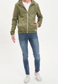 Olivenfarbener leichter Mantel mit Kapuze, Frontreißverschluss, elastischen Bündchen und Saum. Kombiniert mit blauen Slim-Fit-Jeans und weißen Sneakers.