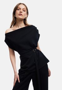 Schwarzes Oberteil mit schulterfreiem Design, Gürteldetail mit einer goldenen herzförmigen Schnalle. Weicher Stoff, lässige Passform, kombiniert mit passenden schwarzen Hosen.