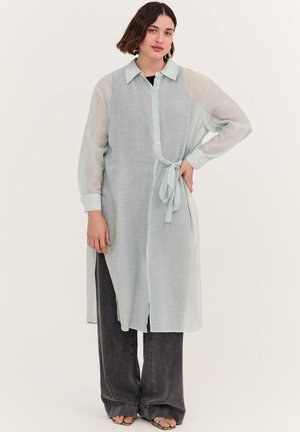 Femme aux cheveux courts et foncés portant une tunique légère et transparente à manches longues boutonnée avec un lien sur le côté, sur un pantalon noir à jambes larges et des sandales.