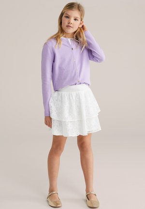 Lila Strickjacke mit Knöpfen, mehrlagiger weißer Spitzrock und goldene Ballerinas. Das Outfit zeichnet sich durch weiche Texturen und kontrastierende Farben aus.