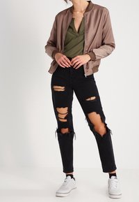 Donna che indossa una giacca bomber color taupe, una maglietta verde oliva a scollo a V, jeans neri strappati e sneaker bianche su uno sfondo semplice.