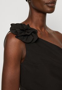 Rochie neagră cu un design cu o singură umăr, accentuată de o floare din țesătură stratificată, mare, pe umăr. Textură fină și formă ajustată.