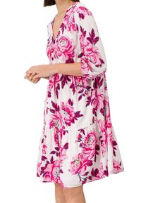 Femme portant une robe blanche ample longueur genoux avec des imprimés floraux rose vif et des manches trois-quarts, debout les mains jointes.