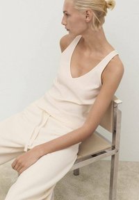 Cremfarbens ärmelloses Oberteil mit tiefem V-Ausschnitt und weichem Stoff, kombiniert mit hellen Cremepant. Model sitzt auf einem schlichten Metallstuhl.