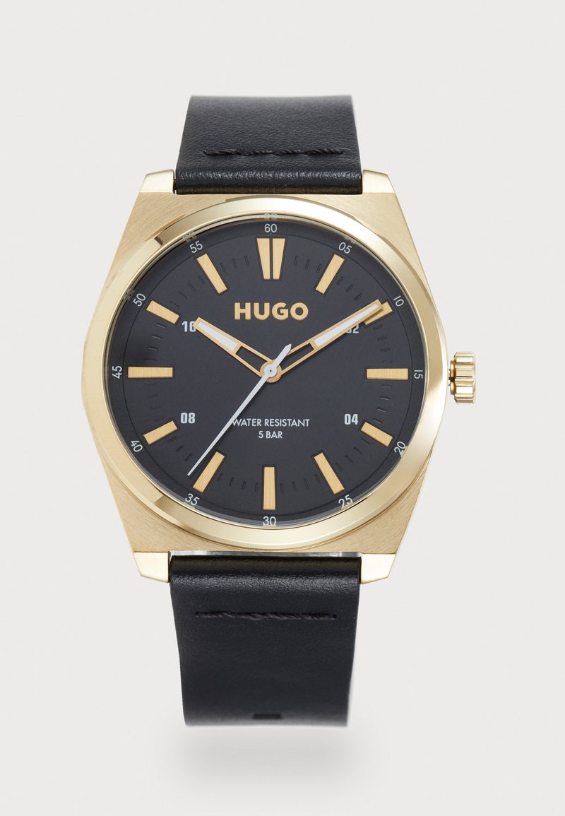 Gold-tone watch with a black dial, hour markers, and leather strap. Features a prominent crown and “HUGO” logo. Water-resistant to 5 bar.