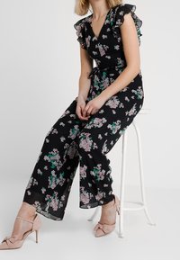 Černý květinový overal s krátkými volánovými rukávy, s růžovými a zelenými květinovými vzory. S uvázaným pasem, volnými nohavicemi a měkkou látkou.