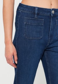Tmavě modré džíny z denimového materiálu s přiléhavým střihem, pěti kapsami a jemným bílým prošíváním podél švů. Hladká textura a klasický vzhled.