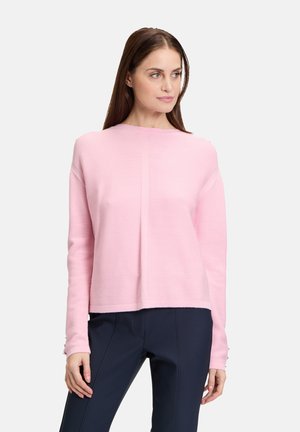 Maglione a coste di un delicato rosa chiaro con scollo rotondo e maniche lunghe, presenta orlo dritto e tessuto texture. Indossato con pantaloni scuri.