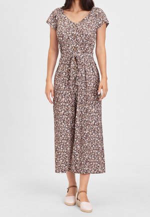 Kobieta ubrana w krótkorękawowy kombinezon w kwiatowy wzór z guzikami z przodu, wiązany w talii, oraz beżowe espadryle z paskiem wokół kostki na białym tle.