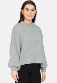 Jonge vrouw met steil bruin haar, goudkleurige oorringen, een loszittende lichtgrijze gebreide trui en zwarte broek, licht naar rechts gericht.