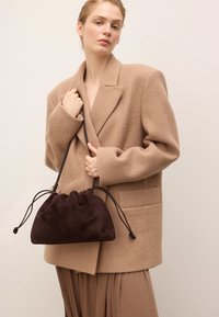 Nainen, jolla on yllään suuri beige takki, pitää tummanruskeaa noblakangasta olevasta nyörilaukusta yksinkertaista taustaa vasten.