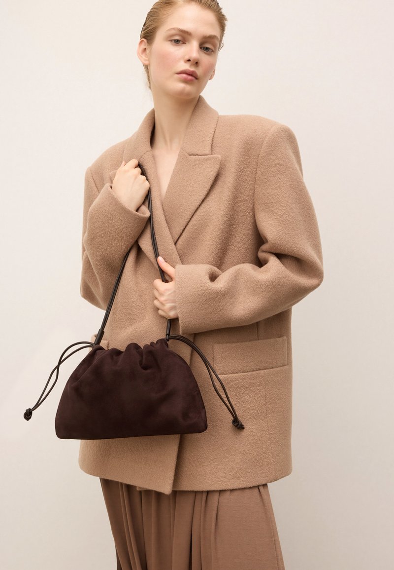 Nainen, jolla on yllään suuri beige takki, pitää tummanruskeaa noblakangasta olevasta nyörilaukusta yksinkertaista taustaa vasten.