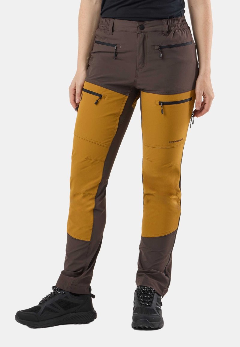 Person wearing brown and mustard outdoor pants with multiple zippered pockets and black hiking shoes on a white background.