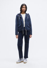 Σκούρο μπλε μπουφάν με κουκούλα, κορδόνια και δύο μπροστινές τσέπες. Φορεμένο πάνω από ένα ανοιχτό πουκάμισο και συνδυασμένο με σκούρο τζιν και γκρίζα παπούτσια.