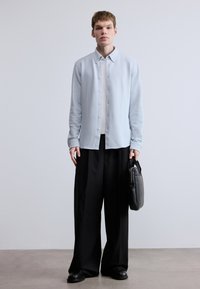 Svetlomodrá košeľa na gombíky s dlhými rukávmi, oblečená cez biele tričko, doplnená čiernymi širokými nohavicami a držiaca čiernu tašku.