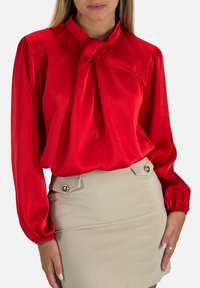 Rød satinbluse med bindebånd i halsen og lange puffede ærmer, kombineret med en beige tætsiddende nederdel med to dekorative knapdetaljer.