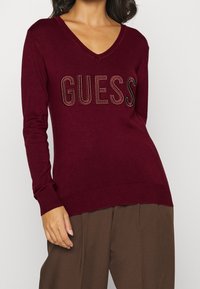 Bordeauxfarbener Strickpullover mit V-Ausschnitt, langen Ärmeln und einem auffälligen "GUESS"-Logo, das mit dekorativen Strasssteinen verziert ist.