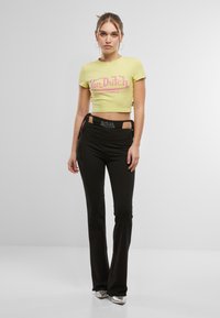 Gul avkortet t-skjorte med rosa "Von Dutch"-logo, sammen med svarte utsvingte bukser som har utskjæringsdetalj og snøring i livet.