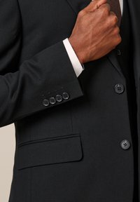 Giacca da uomo nera con finitura strutturata, quattro bottoni neri sul polsino e una tasca a filetto. Polsino della camicia bianca visibile sotto.