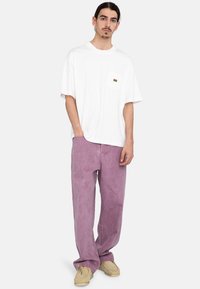 White oversized t-shirt with a chest pocket and multicolor logo, paired with light purple wide-leg pants and beige shoes.