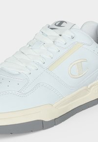 Άσπρο παπούτσι low-top με σόλα σε μπεζ και γκρι, λευκά κορδόνια και ραμμένο λογότυπο "C" στη πλευρική επιφάνεια και στη γλώσσα.