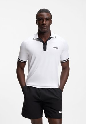 Atletische man die een wit BOSS poloshirt met zwarte bies en zwarte BOSS shorts draagt, staand met handen in de zakken tegen een witte achtergrond.