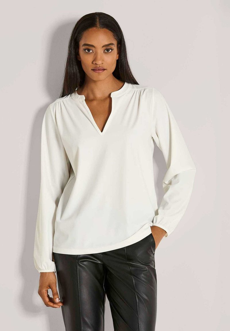Vrouw met lang donker haar, gekleed in een witte blouse met lange mouwen en een V-hals, en zwarte leren broeken, staande tegen een lichte achtergrond.