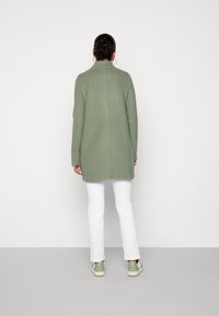 Donna vista da dietro che indossa un cappotto lungo di colore verde salvia, pantaloni bianchi e sneakers verdi e bianchi, in piedi contro una parete bianca uniforme.