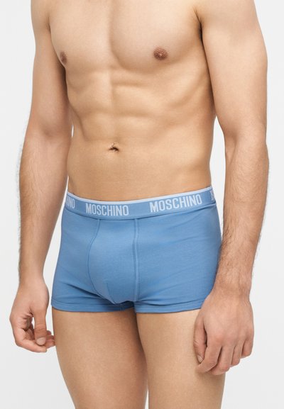 Γυμνασμένος ανδρικός κορμός που φοράει ανοιχτού μπλε boxer Moschino με λογότυπο στη μέση, στέκεται μπροστά σε απλό φωτεινό φόντο.