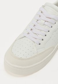 White leather sneaker with a smooth finish, round toe, perforated detailing, flat white laces, and a textured cream rubber sole.