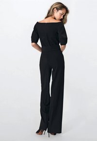 Kobieta z długimi włosami ubrana w czarny kombinezon bez ramion oraz czarne szpilki, stojąca bokiem na jasnym tle.