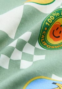 Der grüne Stoff weist ein kariertes Muster mit orangefarbenen und blauen kreisförmigen Grafiken auf, darunter ein lächelndes Gesicht und der Text "100% Kalifornien".