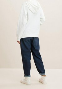 Persona vistiendo una sudadera blanca con capucha, jeans oscuros con puños, y zapatillas altas blancas, de pie frente a un fondo neutro.