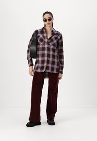 Camicia a quadri rossa e nera con bottoni e due tasche sul petto, abbinata a pantaloni a gamba larga bordeaux e scarpe platform nere. Gli accessori includono occhiali da sole e una borsa nera.