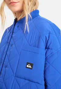 Modrá prošívaná bunda s zapínáním na knoflíky, žebrovaným límcem a náprsným kapsou. Na kapse je logo a materiál je hladký na omak.