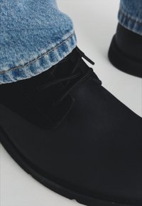 Zapato de cuero negro con un acento de ante y cordones negros, combinado con jeans de denim azul claro que presentan un detalle de dobladillo cosido.