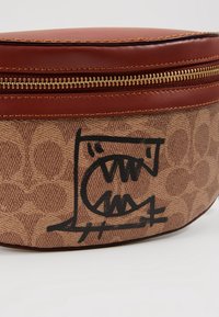 Texturerad brun tygväska med zipp i brons, med en grafik av ett monster med vassa tänder tryckt i svart på sidan.