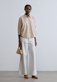 Cămașă bej cu dungi și nasturi, cu mâneci trei sferturi, asortată cu pantaloni largi albi și pantofi cu vârf ascuțit maro. Ține o geantă mică galbenă.