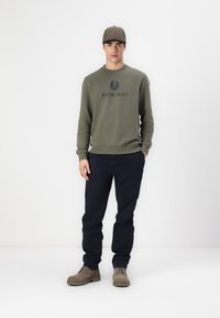 Maslinasto zeleni dukserica s "BELSTAFF" logotipom, mornarske hlače i smeđe čizme na vezivanje. Šarena kapa upotpunjuje odijelo. Jednostavna pozadina.