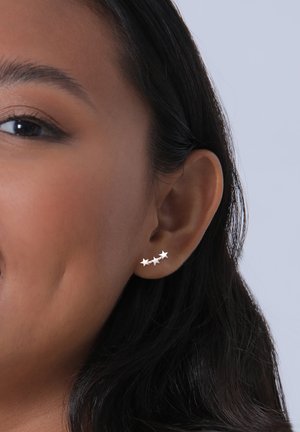 Pendientes en forma de estrella plateada, que presentan tres pequeñas estrellas pulidas dispuestas en un diseño lineal, llevadas en el lóbulo de la oreja.