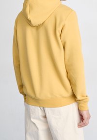 Gul huvtröja med texturerad yta, ribbade muddar och midjeband. Buren med ljusa byxor, sedd bakifrån.