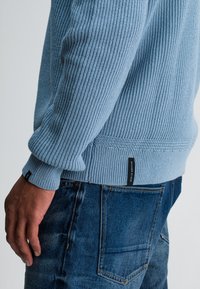 Hellblauer Strickpullover mit geripptem Muster, mit umgeschlagenen Ärmeln und einem kleinen schwarzen Etikett am Saum, kombiniert mit blauen Jeans.