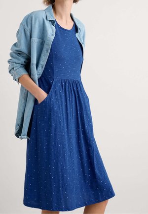 Donna che indossa un vestito blu a pois con tasche e una giacca di jeans chiara sulle spalle, in piedi davanti a uno sfondo uniforme.