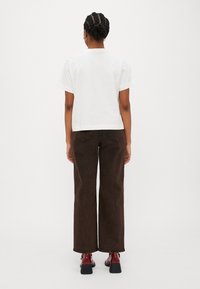 T-shirt bianco a maniche corte in cotone abbinato a pantaloni larghi marroni e scarpe con tacco rosse, visto da dietro su uno sfondo semplice.